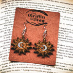 A pair of sunburst-shaped wooden earrings, the "Alchemist's Orbit," featuring a silver-painted crescent moon and star at the center, displayed on a brown "The Griffin and Stag" backing card.