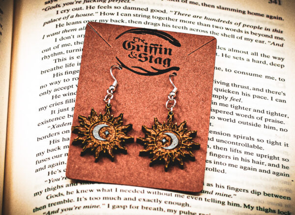 A pair of sunburst-shaped wooden earrings, the "Alchemist's Orbit," featuring a silver-painted crescent moon and star at the center, displayed on a brown "The Griffin and Stag" backing card.