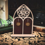 Laser cut and engraved earrings featuring a sun painted gold with a moon and eight-pointed star painted silver in its center hanging from a display stand in the shape of a gothic window in front of stacks of books.