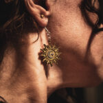 A close-up side profile of a person wearing an Alchemist's Orbit earring, showcasing the detailed sun rays and silver celestial accents against natural sunlight.