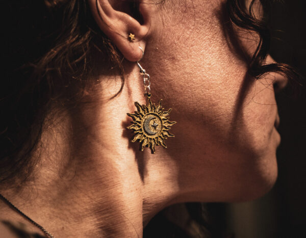 A close-up side profile of a person wearing an Alchemist's Orbit earring, showcasing the detailed sun rays and silver celestial accents against natural sunlight.
