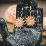 The sunburst earrings displayed on a black, ornate palmistry hand model, surrounded by dark academia-themed decor.
