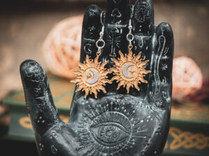 The sunburst earrings displayed on a black, ornate palmistry hand model, surrounded by dark academia-themed decor.