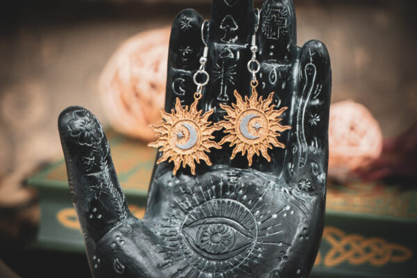 The sunburst earrings displayed on a black, ornate palmistry hand model, surrounded by dark academia-themed decor.