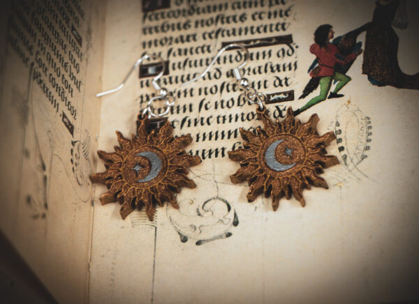 Handcrafted birch wood sun earrings with celestial engravings, styled over an antique manuscript featuring medieval marginalia and whimsical drollery illustrations.