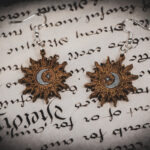 Laser cut and engraved earrings featuring a sun painted gold with a moon and eight-pointed star painted silver in its center on top of a page of calligraphy.