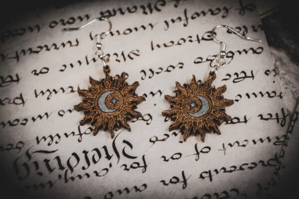 Laser cut and engraved earrings featuring a sun painted gold with a moon and eight-pointed star painted silver in its center on top of a page of calligraphy.