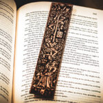 A laser cut and engraved bookmark featuring two men harvesting books from the branches of a tree and tossing them to a woman waiting below on top of the page of an open book.