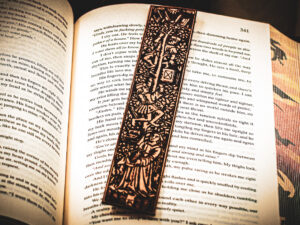 A laser cut and engraved bookmark featuring two men harvesting books from the branches of a tree and tossing them to a woman waiting below on top of the page of an open book.