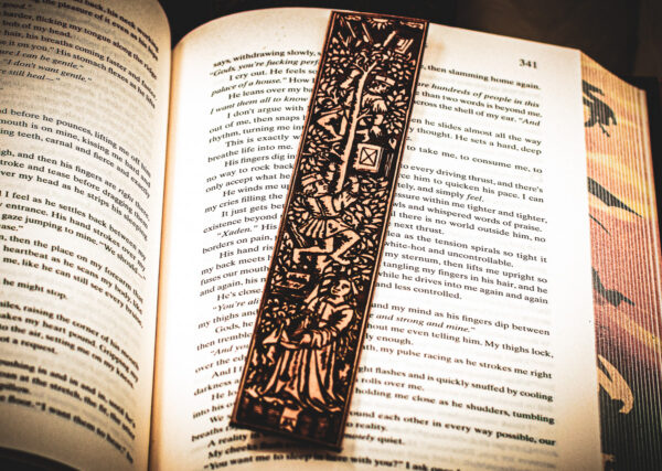 A laser cut and engraved bookmark featuring two men harvesting books from the branches of a tree and tossing them to a woman waiting below on top of the page of an open book.