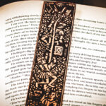 Close-up of the top of a laser cut and engraved bookmark featuring two men harvesting books from the branches of a tree and tossing them to a woman waiting below on top of the page of an open book.