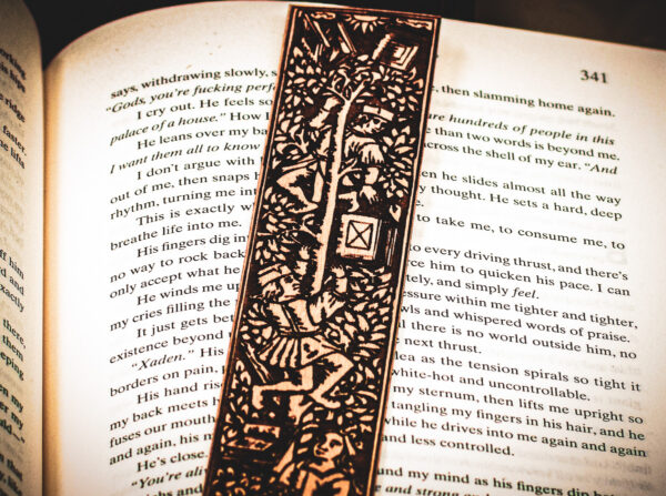 Close-up of the top of a laser cut and engraved bookmark featuring two men harvesting books from the branches of a tree and tossing them to a woman waiting below on top of the page of an open book.