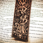 Close-up of the bottom of a laser cut and engraved bookmark featuring two men harvesting books from the branches of a tree and tossing them to a woman waiting below on top of the page of an open book.