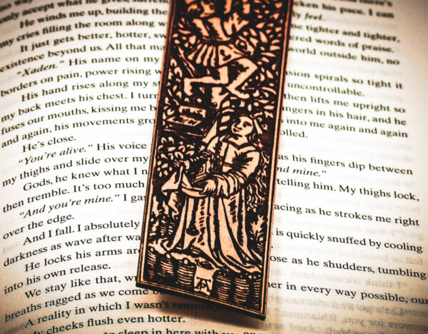 Close-up of the bottom of a laser cut and engraved bookmark featuring two men harvesting books from the branches of a tree and tossing them to a woman waiting below on top of the page of an open book.