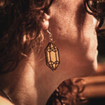 A close-up side profile of a person with curly hair wearing a wooden Arcane Focus earring, highlighting the natural wood grain and delicate gold-toned hardware.