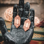 Laser cut and engraved earrings made from raw birch plywood in the shape of crystals hanging from the fingers of a hand sculpture.