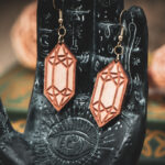 Laser cut and engraved earrings made from raw birch plywood in the shape of crystals hanging from the fingers of a hand sculpture.