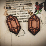 Handcrafted wooden crystal earrings styled atop an open medieval manuscript with whimsical marginalia.