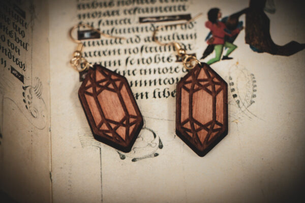 Handcrafted wooden crystal earrings styled atop an open medieval manuscript with whimsical marginalia.