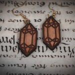 Laser cut and engraved earrings made from raw birch plywood in the shape of crystals on a sheet of calligraphy.