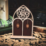 Laser cut and engraved earrings made from raw birch plywood in the shape of crystals hanging from a display in the shape of a gothic window in front of stacks of books.