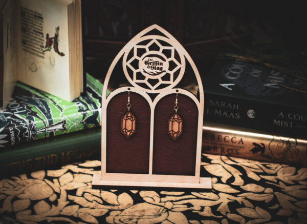 Laser cut and engraved earrings made from raw birch plywood in the shape of crystals hanging from a display in the shape of a gothic window in front of stacks of books.