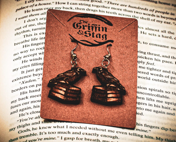 Laser cut and engraved earrings in the shape of a raven perched on a stack of books hanging from a kraft paper card with the Griffin and Stag logo on top of the page of an open book.