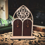 A pair of laser-cut wooden earrings, the "Archivist's Familiar," featuring a raven perched on a stack of books, displayed on a cathedral-style stand with The Griffin and Stag logo.