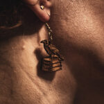 A close-up side profile of a person wearing an Archivist's Familiar earring, highlighting the detailed laser-engraved feathers and book spines against the sunlight.