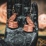 Laser cut and engraved earrings in the shape of a raven perched on a stack of books hanging from the fingers of a hand sculpture.