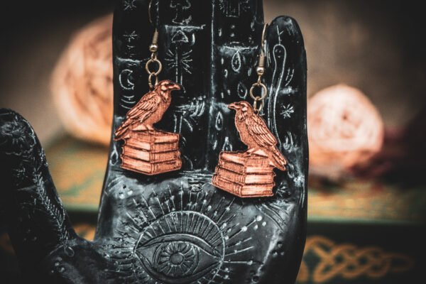 Laser cut and engraved earrings in the shape of a raven perched on a stack of books hanging from the fingers of a hand sculpture.