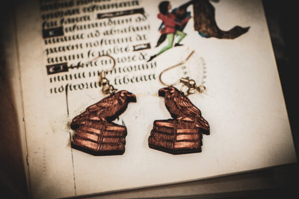Handcrafted birch plywood raven earrings styled over an antique manuscript with medieval marginalia and whimsical drollery illustrations.