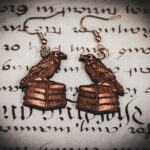 Laser cut and engraved earrings in the shape of a raven perched on a stack of books on top of a page of calligraphy.