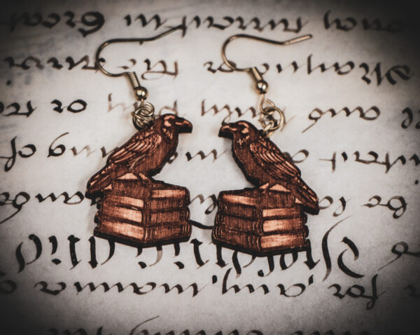 Laser cut and engraved earrings in the shape of a raven perched on a stack of books on top of a page of calligraphy.