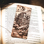 A laser-engraved wooden bookmark featuring a detailed "Astral Sorcerer" wizard with a long beard and star-patterned hat, resting on the open pages of a fantasy novel.