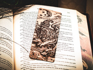 A laser-engraved wooden bookmark featuring a detailed "Astral Sorcerer" wizard with a long beard and star-patterned hat, resting on the open pages of a fantasy novel.