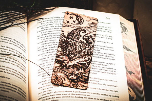 A laser-engraved wooden bookmark featuring a detailed "Astral Sorcerer" wizard with a long beard and star-patterned hat, resting on the open pages of a fantasy novel.