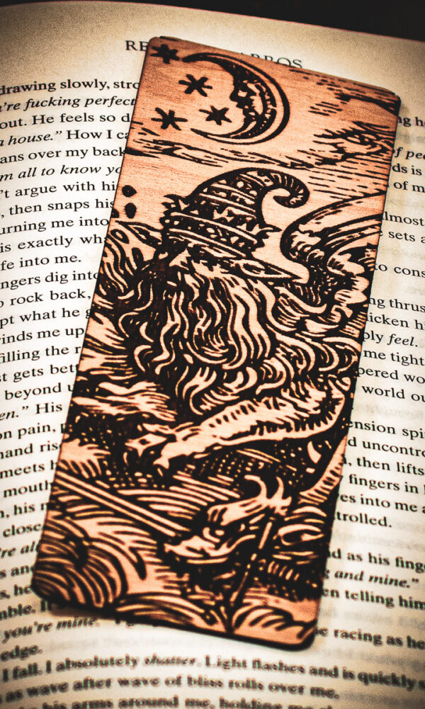 A detailed view of a handcrafted birch wood bookmark depicting a sorcerer beneath a crescent moon and stars, highlighting the intricate wood grain and dark engraved lines.