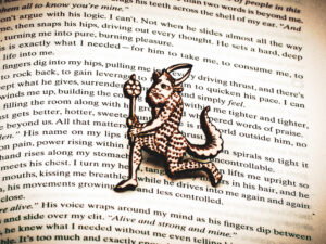 A laser engraved birch plywood pin featuring a running satyr holding a branch from the Aberdeen Bestiary on the page of an open book.