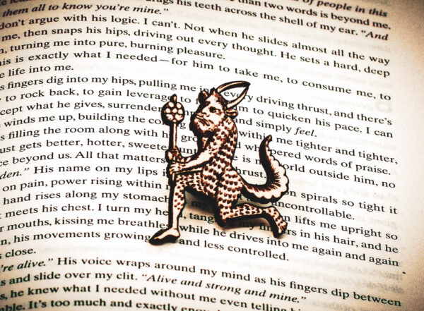 A laser engraved birch plywood pin featuring a running satyr holding a branch from the Aberdeen Bestiary on the page of an open book.