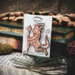 A laser cut and engraved magnet made from raw birch plywood in the shape of a satyr holding a branch from the Aberdeen Bestiary in a cellophane bag with a gray card background with stylized trees leaning against a stack of books.
