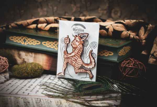 A laser cut and engraved magnet made from raw birch plywood in the shape of a satyr holding a branch from the Aberdeen Bestiary in a cellophane bag with a gray card background with stylized trees leaning against a stack of books.