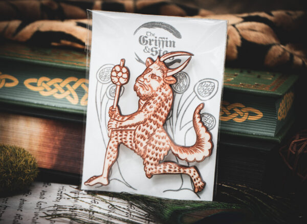 A laser cut and engraved magnet made from raw birch plywood in the shape of a satyr holding a branch from the Aberdeen Bestiary in a cellophane bag with a gray card background with stylized trees leaning against a stack of books.