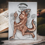 A laser cut and engraved magnet made from raw birch plywood in the shape of a satyr holding a branch from the Aberdeen Bestiary in a cellophane bag with a gray card background with stylized trees leaning against an open book.
