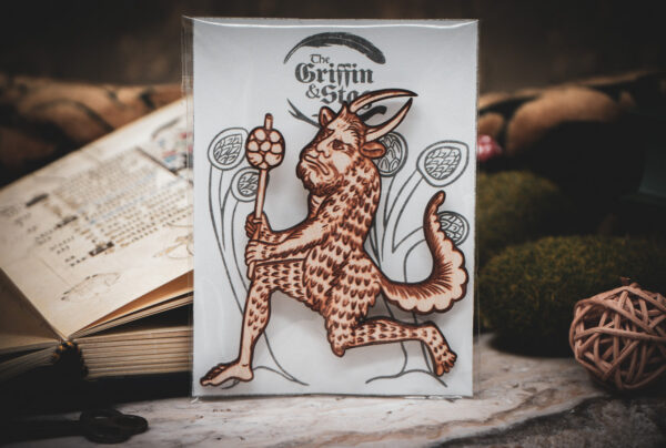 A laser cut and engraved magnet made from raw birch plywood in the shape of a satyr holding a branch from the Aberdeen Bestiary in a cellophane bag with a gray card background with stylized trees leaning against an open book.