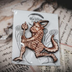 A laser cut and engraved magnet made from raw birch plywood in the shape of a satyr holding a branch from the Aberdeen Bestiary in a cellophane bag with a gray card background with stylized trees on top of various papers on a stone surface.