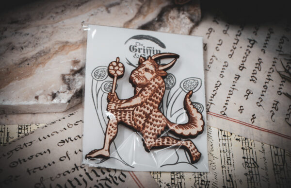 A laser cut and engraved magnet made from raw birch plywood in the shape of a satyr holding a branch from the Aberdeen Bestiary in a cellophane bag with a gray card background with stylized trees on top of various papers on a stone surface.