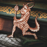 A laser cut and engraved magnet made from raw birch plywood in the shape of a satyr holding a branch from the Aberdeen Bestiary leaning against a stack of books.