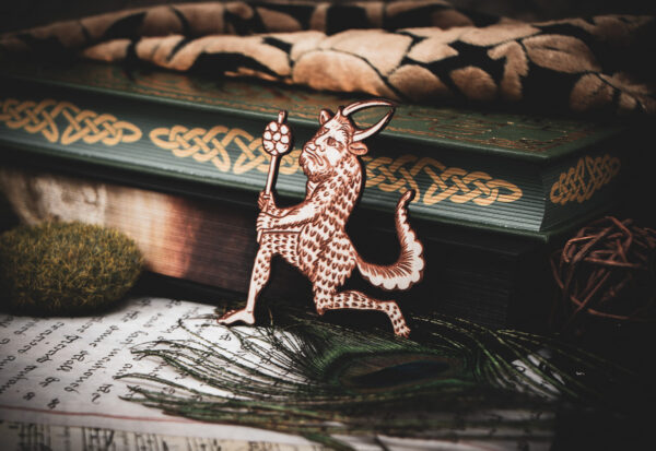 A laser cut and engraved magnet made from raw birch plywood in the shape of a satyr holding a branch from the Aberdeen Bestiary leaning against a stack of books.
