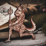 A laser cut and engraved magnet made from raw birch plywood in the shape of a satyr holding a branch from the Aberdeen Bestiary leaning against an open book.